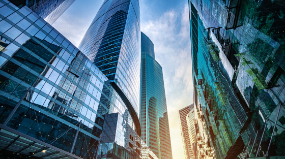 An upward view of large glass enclosed skyscrapers with blue tones and sunlight subtly rising between the buildings