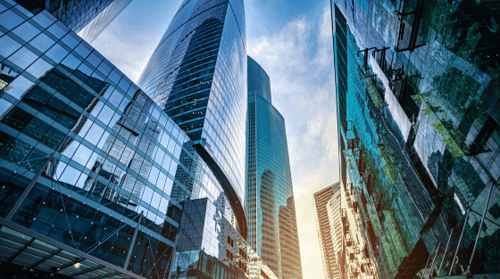 An upward view of large glass enclosed skyscrapers with blue tones and sunlight subtly rising between the buildings